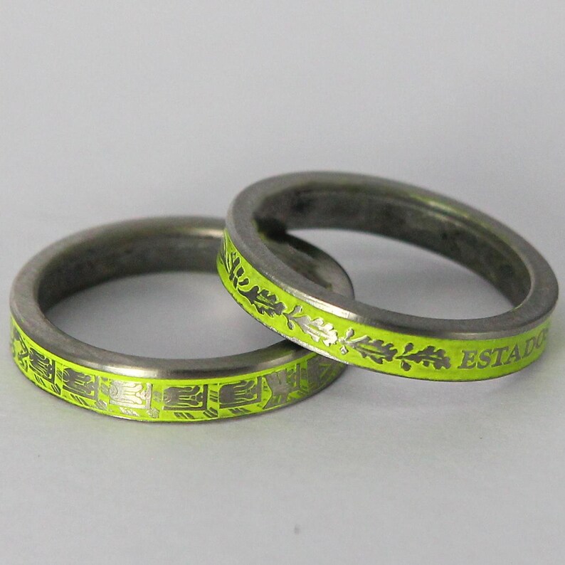 K&ouml;nnte beinhalten: Zwei silberne Ringe mit einer leuchtend gelben Emaille-Oberfl&auml;che. Ein Ring hat ein sich wiederholendes Muster aus stilisierten Bl&auml;ttern und dem Text "ESTADIO". Der andere Ring hat ein sich wiederholendes Muster aus stilisierten geometrischen Formen.