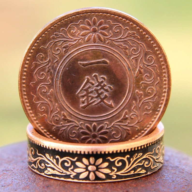 May include: A copper-colored coin with Japanese characters and floral detailing rests atop a black and gold ring with a floral design. The coin and ring are set against a blurred green and yellow background.