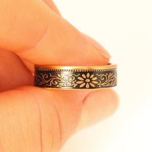 May include: A gold-toned ring with a black band featuring a floral design. The ring's band is adorned with a repeating pattern of flowers and swirling vines. The ring is held between fingers against a white background.
