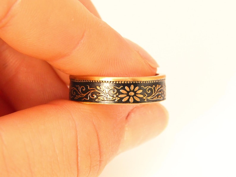 May include: A gold-toned ring with a black band featuring a floral design. The ring's band is adorned with a repeating pattern of flowers and swirling vines. The ring is held between fingers against a white background.