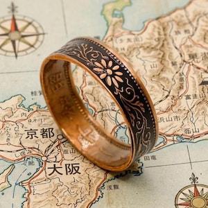 May include: A copper-toned ring with a black floral design band, set against a vintage map of Japan. The ring's band features a repeating pattern of flowers and leaves. The map includes Japanese text and a compass rose.