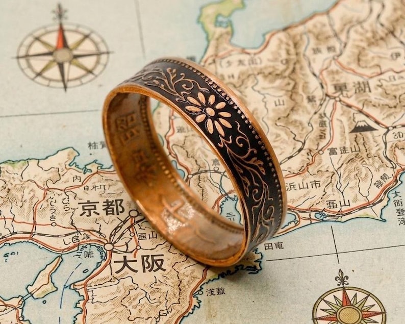 May include: A copper-toned ring with a black floral design band, set against a vintage map of Japan. The ring's band features a repeating pattern of flowers and leaves. The map includes Japanese text and a compass rose.