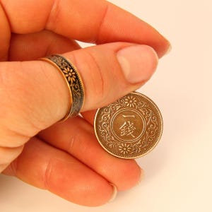 May include: A gold-toned ring with a black floral design is worn on a finger. A vintage coin with Chinese characters and floral details is held in the same hand. The coin has a circular design with a central inscription.