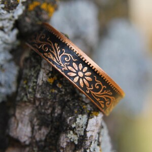 May include: A copper-colored metal bracelet with a black center band. The band features a floral design with a daisy in the center. The bracelet is resting on a tree branch.