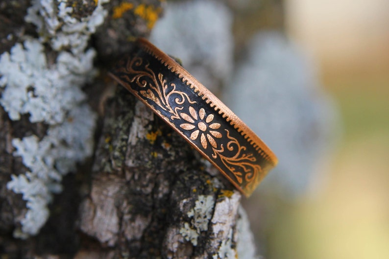 May include: A copper-colored metal bracelet with a black center band. The band features a floral design with a daisy in the center. The bracelet is resting on a tree branch.