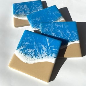 May include: Set of four square coasters with a beach scene. The coasters feature a blue ocean with white foam waves crashing on a tan sandy beach.