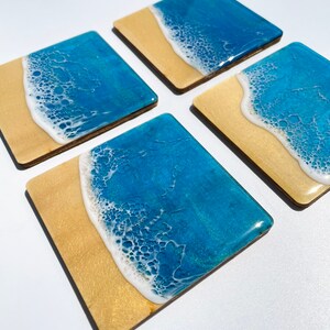 May include: Set of four square coasters with a resin design of a beach scene. The coasters feature a blue ocean with white foam waves crashing on a gold sand beach.