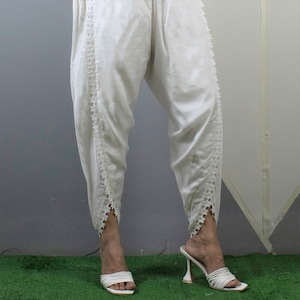 May include: Off-white, satin-like pants with a unique, gathered design. The pants feature delicate, floral-shaped lace trim along the outer seams. The model is wearing white, strappy high-heeled sandals. The background is a mix of gray and green.