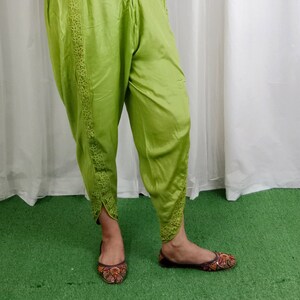 May include: Green pants with lace trim and a fitted silhouette. The pants have a slight flare at the bottom and are worn with brown shoes.