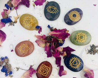 May include: Seven smooth, oval-shaped stones with gold chakra symbols. The stones are in various colours, including green, purple, brown, and grey. The stones are arranged on a white surface with dried flower petals.