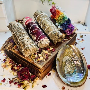 Può includere: Una scatola di legno piena di diversi bastoncini di incenso, tra cui salvia bianca, rosa e mazzi di fiori arcobaleno. Una grande conchiglia di abalone è in primo piano.