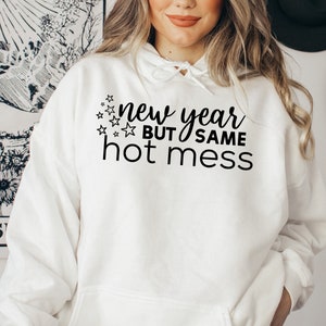 May include: White hoodie with black text that reads "new year but same hot mess" with a scattering of stars around the text.