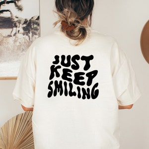 May include: A white t-shirt with a black graphic that says "JUST KEEP SMILING" in a retro, bubbly font.