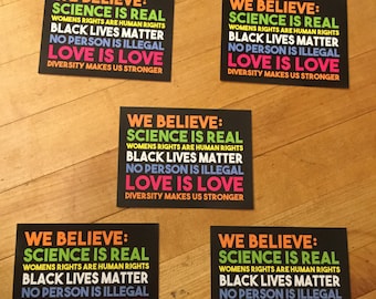 May include: Four black signs with white text and colorful accents. The signs read "We Believe: Science is Real, Women's Rights are Human Rights, Black Lives Matter, No Person is Illegal, Love is Love, Diversity Makes Us Stronger."