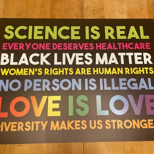 May include: A black and white sign with the text "SCIENCE IS REAL", "EVERYONE DESERVES HEALTHCARE", "BLACK LIVES MATTER", "WOMEN'S RIGHTS ARE HUMAN RIGHTS", "NO PERSON IS ILLEGAL", "LOVE IS LOVE", and "DIVERSITY MAKES US STRONGER".