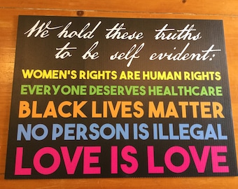 May include: A black sign with white text that reads "We hold these truths to be self evident:" followed by five lines of text in different colours: "WOMEN'S RIGHTS ARE HUMAN RIGHTS", "EVERYONE DESERVES HEALTHCARE", "BLACK LIVES MATTER", "NO PERSON IS ILLEGAL", and "LOVE IS LOVE".