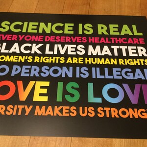 May include: A black and white poster with the text "SCIENCE IS REAL", "EVERYONE DESERVES HEALTHCARE", "BLACK LIVES MATTER", "WOMEN'S RIGHTS ARE HUMAN RIGHTS", "NO PERSON IS ILLEGAL", "LOVE IS LOVE", and "DIVERSITY MAKES US STRONGER".