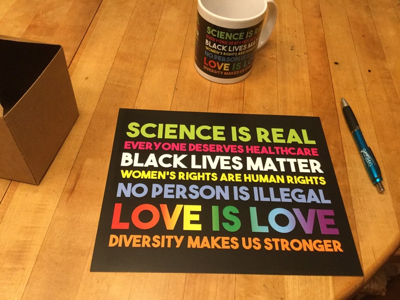 May include: A black and white poster with the text "SCIENCE IS REAL", "EVERYONE DESERVES HEALTHCARE", "BLACK LIVES MATTER", "WOMEN'S RIGHTS ARE HUMAN RIGHTS", "NO PERSON IS ILLEGAL", "LOVE IS LOVE", and "DIVERSITY MAKES US STRONGER" in different colours.