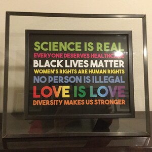 May include: A framed print with a black background and colourful text that reads "SCIENCE IS REAL", "EVERYONE DESERVES HEALTHCARE", "BLACK LIVES MATTER", "WOMEN'S RIGHTS ARE HUMAN RIGHTS", "NO PERSON IS ILLEGAL", "LOVE IS LOVE", and "DIVERSITY MAKES US STRONGER".