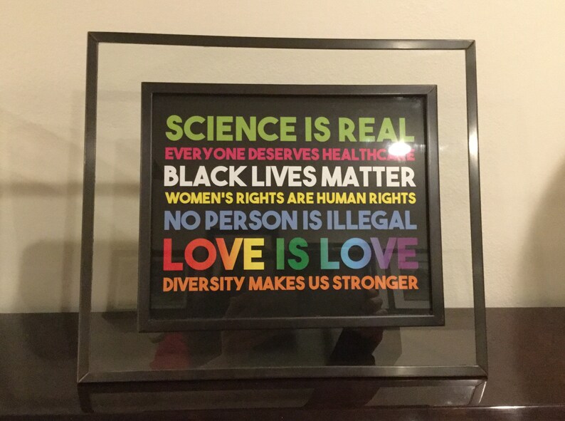 May include: A framed print with a black background and colourful text that reads "SCIENCE IS REAL", "EVERYONE DESERVES HEALTHCARE", "BLACK LIVES MATTER", "WOMEN'S RIGHTS ARE HUMAN RIGHTS", "NO PERSON IS ILLEGAL", "LOVE IS LOVE", and "DIVERSITY MAKES US STRONGER".