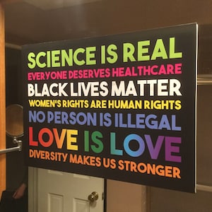 May include: A black sign with white text and rainbow lettering. The text reads "Science is real, Everyone deserves healthcare, Black lives matter, Women's rights are human rights, No person is illegal, Love is love, Diversity makes us stronger."