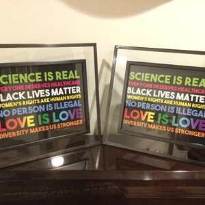 May include: Two framed prints with a black background and rainbow coloured text. The text reads "Science is real, Everyone deserves healthcare, Black lives matter, Women's rights are human rights, No person is illegal, Love is love, Diversity makes us stronger."