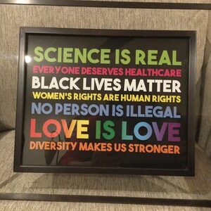 May include: Framed print with a black background and colourful text that reads "Science is real", "Everyone deserves healthcare", "Black lives matter", "Women's rights are human rights", "No person is illegal", "Love is love", and "Diversity makes us stronger".