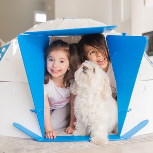Puede incluir: Una casita de juegos blanca y azul con dos niños y un perro pequeño blanco dentro. La casita está hecha de paneles blancos con detalles azules. Los niños sonríen y miran por la abertura de la casita.