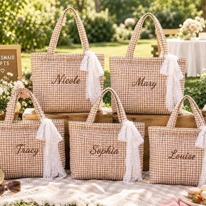 May include: Five quilted tote bags in a tan and white gingham pattern, each featuring a name embroidered in brown script and a white lace ribbon tied to the handle. The names Nicole, Mary, Tracy, Sophia, and Louise are displayed on the bags.