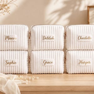 May include: Six white rectangular cosmetic bags with a vertical ribbed texture and gold zippers. Each bag features a central white rectangular patch with a name embroidered in gold script: Maeve, Delilah, Charlotte, Sophie, Grace, and Harper.