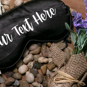 May include: Black satin sleep mask with the text "Your Text Here" in white lettering. The mask is surrounded by small stones and a bouquet of lavender.