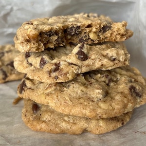May include: A stack of four chocolate chip cookies on white parchment paper. The top cookie is broken in half, revealing the chocolate chips inside.
