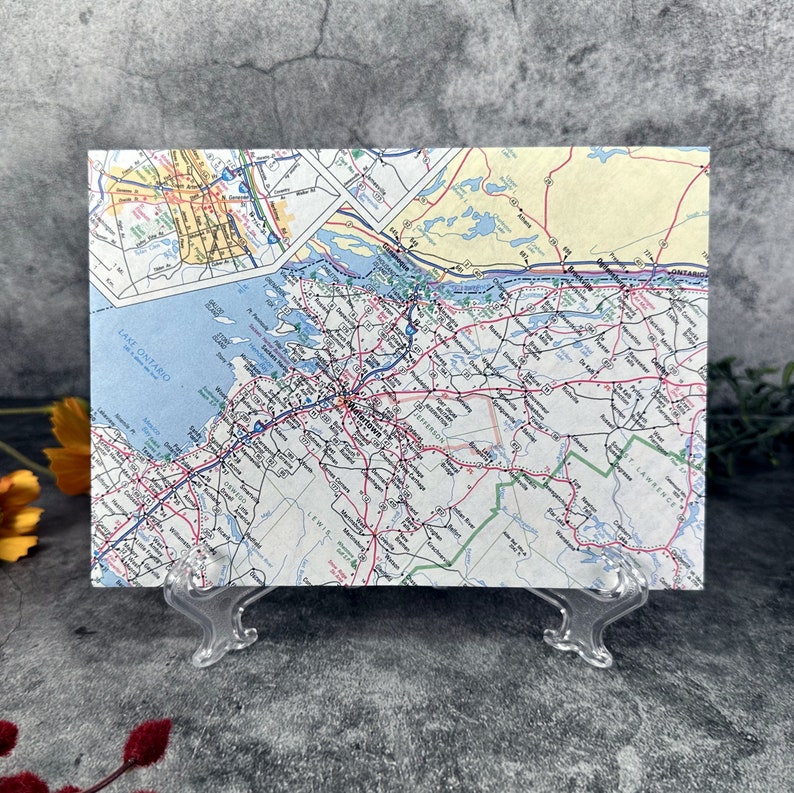 May include: A vintage map of the northeastern United States, showing the state of New York and surrounding areas. The map is printed on paper and has a light blue, yellow, and green color scheme. The map is displayed on a clear plastic stand.