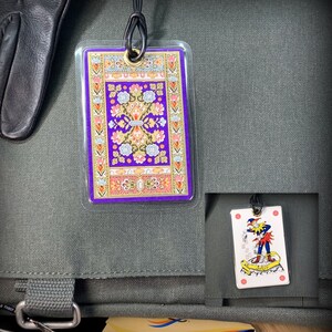 May include: A green canvas bag with a purple and gold patterned fabric tag attached to it. The tag has a cord with a metal loop. A playing card with a jester on it is attached to the bag with a string.