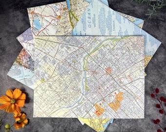 May include: A stack of vintage maps with a detailed city map on top. The maps are in various shades of blue, green, and yellow. The city map shows streets, roads, and landmarks. The text on the maps includes "Ocean", "Tasman Sea", and "Pacific Sea".