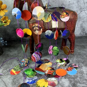 May include: A colorful mobile featuring circular cutouts with various images, strung together with purple thread. The mobile hangs in front of a wooden horse statue. The cutouts display images of flowers, landscapes, and abstract art.