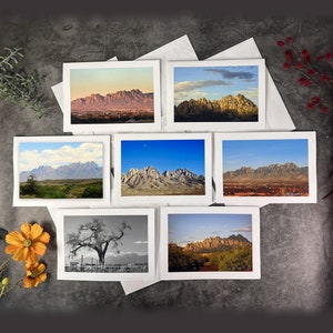 May include: A set of eight greeting cards featuring different scenic views of a mountain range. The cards are white with a white border and are arranged in two rows of four. The cards are all the same size and shape.