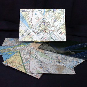 May include: A collection of folded maps, some with road maps and some with city maps, are stacked on top of each other. The maps are all different sizes and colors. The maps are on a black surface.