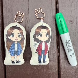 以下が含まれることがあります： チェック柄のシャツとショートパンツまたはジーンズを履いた漫画のキャラクターが特徴の2つのぬいぐるみキーホルダー。キーホルダーには、ローズゴールドのウサギ型の留め金が付いています。緑色のSharpieマーカーがキーホルダーの隣にあります。