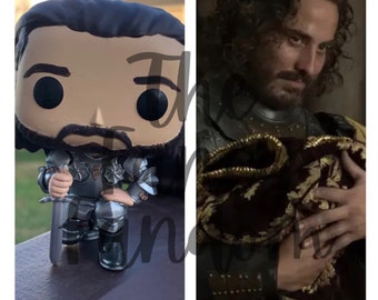 Può includere: Una statuetta in vinile pop di un uomo con la barba e i capelli lunghi, che indossa un'armatura argentata e tiene una spada. È seduto su una superficie di legno. La statuetta è accanto a una foto di un uomo con la barba e i capelli lunghi, che indossa una tunica marrone scuro e tiene un tessuto rosso e dorato.