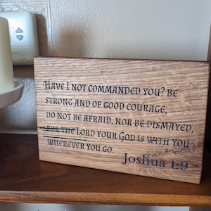 May include: Wooden plaque with engraved text from Joshua 1:9. The plaque is rectangular, with a natural wood grain finish. The text is in a classic font and reads, "Have I not commanded you? Be strong and of good courage..."