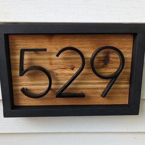 May include: A rectangular house number sign featuring the digits 529 in a bold, black, sans-serif font. The numbers are mounted onto a natural, light-brown wood grain panel, which is enclosed within a solid, matte black rectangular frame.