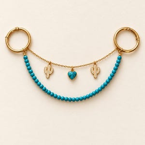 Puede incluir: Un accesorio de cadena en tono dorado con dos cierres circulares conectados por una hilera de pequeñas cuentas turquesas y una fina cadena dorada. Tres dijes cuelgan de la cadena: dos siluetas de cactus doradas y un corazón turquesa.