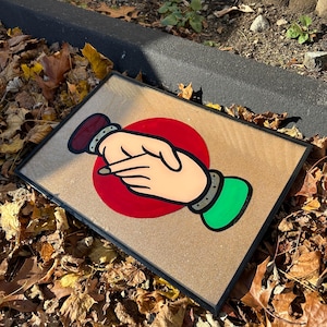 May include: A framed artwork featuring a stylized illustration of two hands shaking, set against a red circle. The hands are depicted with red and green wristbands, and the background is a light brown color. The frame is black.