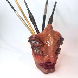 Könnte beinhalten: Ein Keramik-Pinselhalter in Form eines Gesichts mit vier Augen, einer großen Nase und einem roten Mund. Der Halter ist in Braun-, Rot- und Weißtönen bemalt.