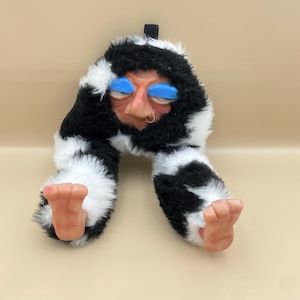 May include: A whimsical plush figure with a face, blue eyelids, and a nose ring. The figure has black and white fur and two feet. The feet are a flesh tone. A black loop is attached to the top of the figure.