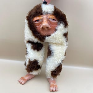 May include: A whimsical, handmade art piece featuring a human-like face with a realistic nose and eyes, integrated into a brown and white patterned furry hat and pants. The piece includes two small feet. The art is held up by a hand.
