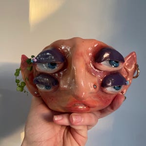 Puede incluir: Una escultura de cerámica hecha a mano de una cara con múltiples ojos azules y párpados morados. La escultura tiene una tez rosada-melocotón, con pequeñas plantas verdes y cuentas que adornan una oreja. La escultura se sostiene en una mano.