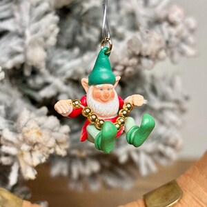 May include: A Christmas ornament featuring a smiling elf with a white beard, wearing a green hat, red jacket, and green shoes. The elf is holding a gold beaded necklace. The ornament is suspended by a silver wire.