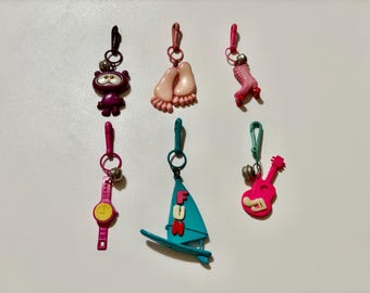 Dijes de plástico retro vintage de los años 80: pies descalzos, patín, reloj rosa, windsurfista, guitarra rosa y figura de ojos saltones morados.